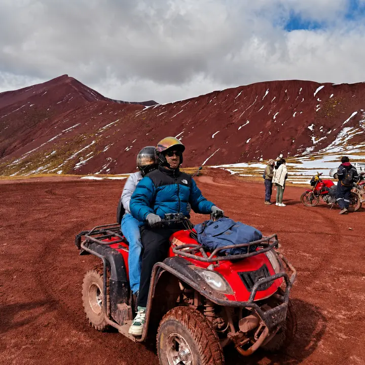 Two riders crossing the Red Valley on an ATV beneath snow-streaked slopes.