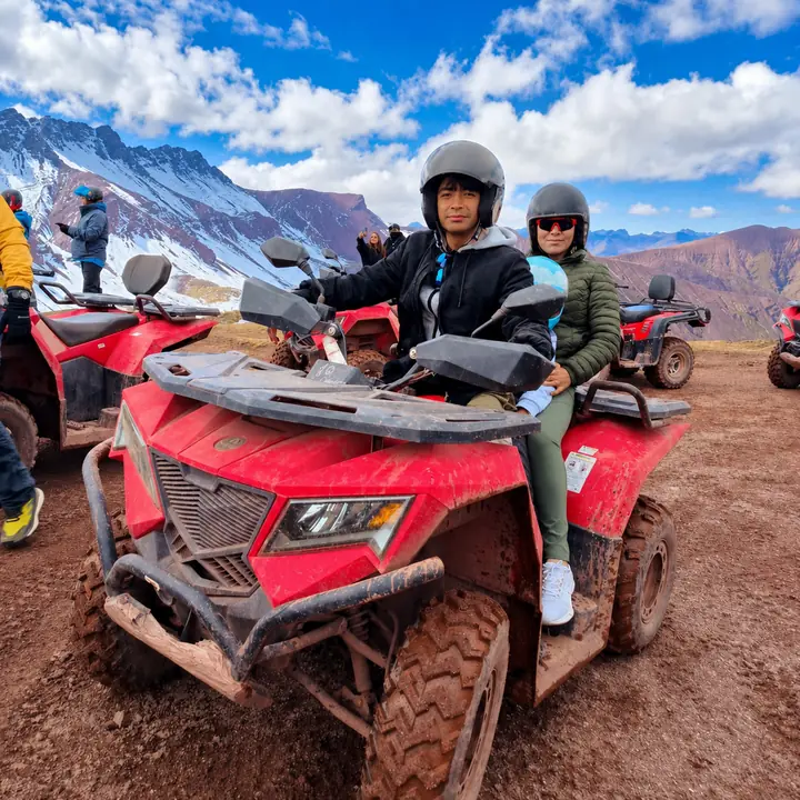 Two travelers seated on a red ATV with snow-covered peaks behind them.