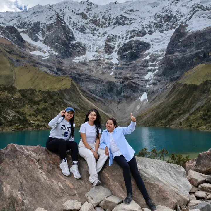 Three women sitting on rocks above Humantay Lake and snowy mountains.