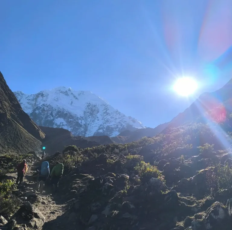 Trekkers climbing a rocky Andean trail toward Salkantay Mountain under the bright morning sun.